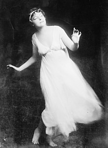 A white woman wearing a white flowing dress, dancing against a dark background