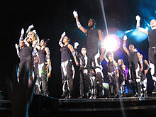 Левый профиль группы людей, прыгающих на сцену с вытянутой вверх правой рукой. На них черные топы, брюки в черно-белую полоску и браслеты. На правой руке у них белые перчатки. Сверху они подсвечиваются белым светом.