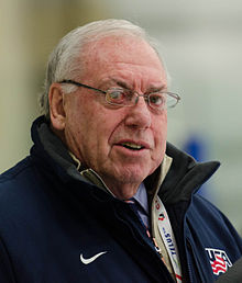 Elderly man with white hair and eyeglasses, wearing a navy blue jacket with a USA Hockey logo