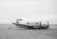 A twin-engined World War II-era monoplane lying on its belly on grass. Both of its propellers are twisted and another aircraft is flying near the ground behind it.