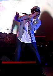 Мужчина на сцене с микрофоном в куртке с капюшоном, белой рубашке и синих джинсах.