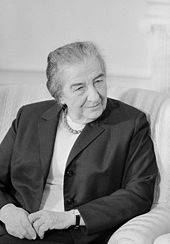 A dignified woman in her seventies sitting on a chair with her hands placed on her lap, staring intently to one side as if speaking to someone out of frame.