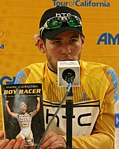 Кавендиш сидит перед микрофоном с копией своей книги Boy Racer