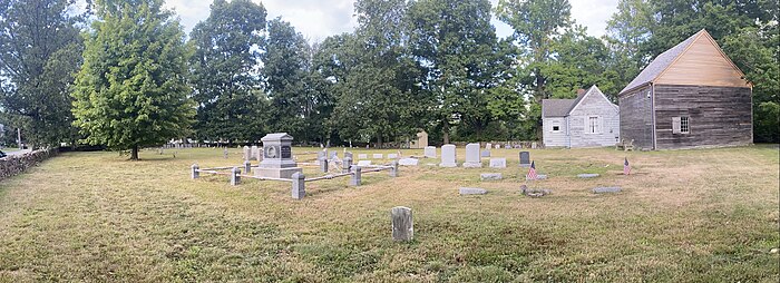 A Quaker cemetery including green grass and shady trees stands under a partly cloudy sky. The gravestones are old and faded; many are crooked in the ground: A few American flags wave above the gravestones. Two houses are on the right; one is a small white house, built within the last 150 years, presumably to care for the property, and the other is the original historical Quaker meeting house. It is wood from bottom to top, although the triangle on the wall forming the roof is lighter than the rest due to restoration.