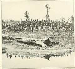 Sepia-tone photo shows about 50 soldiers standing at attention with a pool of water and a fallen tree in the foreground.