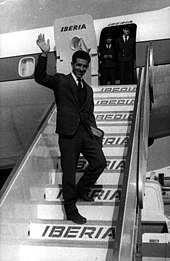 Photographie en noir et blanc d'un homme en costume saluant la foule dans les marches pour accéder à un avion de la compagnie Iberia.