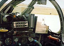 Inside view of a cramped cockpit, facing forward and slightly to the right. Metal windscreen support frames and a gun sight partially block the view outside.