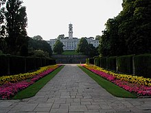 A white building topped by a tower, at the end of a flowery driveway.