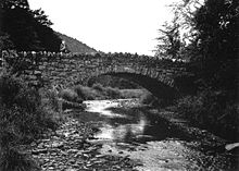 Black and white photograph of the side of a stone bridge arching over a shallow rocky stream. The torso and head of a person wearing a hard hat can be seen on the left side of the bridge.