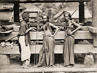 Tri batakaj militistoj kun lancoj kaj spadoj antaŭ ligna konstruaĵo en Indonezio (ĉ. 1870)