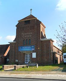 Three-quarter view of a brown-brick church with a short, stubby tower in the foreground. This is topped with a shallow spire and a crucifix. There is a concrete bench on a grassed area in front of the building.