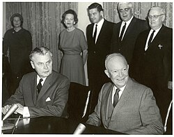 Diefenbaker et un homme chauve souriant sont assis à une table. Deux femmes et trois hommes se tiennent derrière eux.