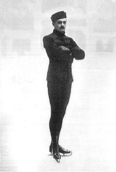 A male figure skater poses with crossed arms for a shot at an indoor ice rink.