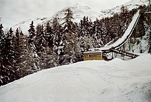 Image d'un tremplin de saut à ski avec en bas l'inscription "St. Moritz" et derrière lui, des arbres enneigées.