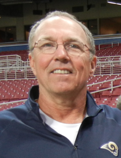 Posed head and shoulders photograph of Boudreau wearing eyeglasses and blue pullover bearing a St. Louis Rams logo standing in an apparently empty stadium