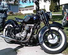 A mildly customized Sunbeam S7 motorcycle sits along the edge of a driveway. It shows evidence of normal wear and tear, but appears to be in very good condition. It's all black, with a vaguely egg-plant shaped fuel tank that features rubber pads for the rider's knees, and white-wall tires.