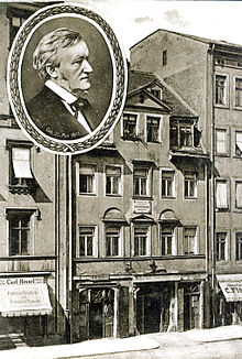 A postcard of a five-storey building with shops on the ground floor and garret windows in the roof. A round inset has a picture of Wagner in middle age.