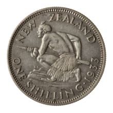 A coin featuring a crouched Māori atop a rocky escarpment. He holds a taiaha war-staff with both hands pointing it and facing forward off the escarpment. He is muscular and shirtless, but wears a piupiu skirt.