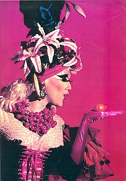 Een dragqueen in paarse kleding met een hoofdtooi met bloemen possert.