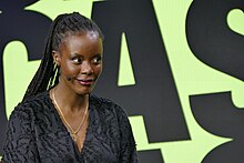 Bild (Oberkörper) einer schwarzen Frau mit Cronrow-Frisur, die ein Kopfbügelmikrofon trägt. Sie lächelt leicht und blickt nach rechts. Hinter ihr eine gelbe Wand mit großen schwarnzen Buchstaben, C, A, und S sind erkennbar.