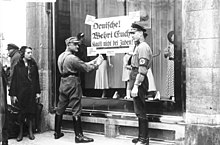 SA paramilitaries outside a Berlin store on 1 April 1933 during the Nazi boycott of Jewish businesses. The sign reads: "Germans! Defend yourselves! Don't buy from Jews!" Bundesarchiv Bild 102-14468, Berlin, NS-Boykott gegen judische Geschafte.jpg