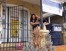 A woman standing on the patio of a yellow-painted brick row house. A sign in the door reads "Casa Ruby".