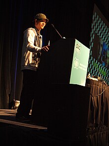 A 25-year-old Swedish man in a grey jacket, black pants, and green hat speaking at a conference.