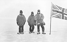 Three men in heavy clothing stand in line on an icy surface, next to a flagstaff from which flies the flag of the United Kingdom of Great Britain and Ireland