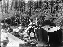 photo en noir et blanc de deux hommes debout dans une forêt, l'un prêt à fendre un morceau de tronc, l'autre mettant en forme une douelle avec une plane.