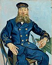 a long-bearded man with a blue uniform and hat is seated in a chair facing forward with his right arm on the chair's arm and left arm on a table and with a pastel blue background