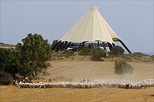 A large sand-coloured conical tent on a small hill before a pale blue sky, with a few trees and a herd of sheep on dry grassland in the foreground