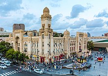 Zdjęcie. W centralnej części widoczny w rzucie izometrycznym budynek dworca. Przed nim plac z kilkudziesięcioma poruszającymi się ludźmi, kioskiem, zejściem na stację metra. Po lewej stronie zdjęcia przed budynkiem kilkupasmowa ulica z samochodami zatrzymanymi przed widocznym u dołu fotografii przejściem dla pieszych, odgrodzona od budynku szpalerem drzew. W tle niebo z chmurami. Budynek wykonany z kamienia o barwie jasnego piasku, posiadający trzy wysokie piętra z licznymi oknami. Bryła prostopadłościenna z licznymi zdobieniami. W połowie każdej ściany wywyższony ponad bryłę budynku prostokątny portal z prowadzącymi na wysokość pierwszego piętra szerokimi schodami. W rogu budynku, widocznym w środkowej części fotografii, ozdobna wieża na planie kwadratu, o obłym zakończeniu, około dwukrotnie wyższa od bryły budynku. W połowie odległości pomiędzy narożnikami budynku a portalami umieszczone ozdobne wieżyczki na planie kwadratu, wysunięte nieco poza bryłę budynku, o wysokości portalu, o obłym zakończeniu.