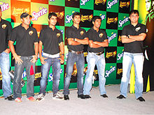 A group of men standing, wearing black T-shirts and blue faded jeans. All of them are looking to the left of the image. The backdrop has alternate black and green boxes.