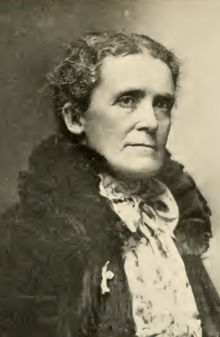 An older white woman with a stern expression, with grey hair parted center and dressed to the nape; she is wearing a jacket with a high fur collar, and a white ribbon for temperance is visible on her lapel