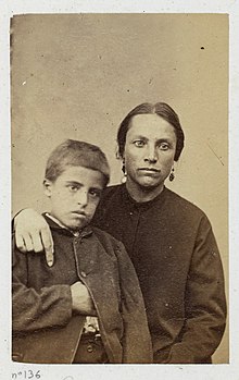 Portrait en photographie sépia d'une femme habillée simplement, au visage rehaussé de deux boucles d'oreille, et posant son bras droit sur l'épaule de son fils, au regard hagard.