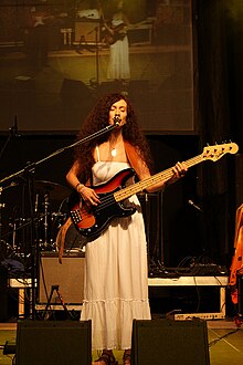 Slim woman with long dark hair wearing light coloured dress at a microphone on stage playing an electric bass guitar under spotlighting