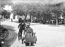 A photograph from 1899 depicting a woman using a wheeled chair, with a man standing behind as attendant; they are outdoors in a wide unpaved path.