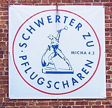 An einer roten Ziegelwand hängt ein Banner mit dem kreisrunden Symbol der Friedensbewegung „Schwerter zu Pflugscharen“. Es ist rot umrandet. Im Kreis steht oben entlang des Randes „Schwerter zu“ und unten „Pflugscharen“. In der Mitte ist die gleichnamige Bronzeskulptur abgebilder.