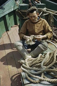 Durant la Seconde Guerre mondiale, à Brixham dans le Devon, sur le pont d’un bateau de pêche, un marin-pêcheur belge qui a gagné l’Angleterre répare un bout usagé.