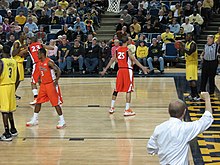 a man in a white shirt makes a signal to basketball players on the court with his fist in the air from the sidelines. He is viewed from behind.