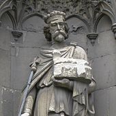 Statue en pierre grise d'un homme barbu et couronné qui tient un sceptre dans la main droite et une église en modèle réduit dans la main gauche.