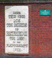 Photographie d'un mur de briques rouges. Sur la gauche, une grande plaque blanche porte une inscription en caractères ouvragés. Une petite plaque verte est fixée à côté.