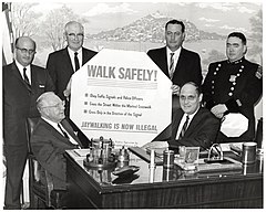Des hommes politiques entourant un panneau sur lequel est notamment écrit « Walk safely ! Jaywalking is now illegal ».