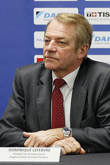 Homme en costume de trois quarts, cheveux gris, bras croisés, assis à une table lors d'une conférence de presse sportive.