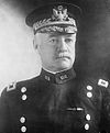 Man facing forward in dress uniform with two vertical columns of buttons and hat with gold trim