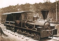 An old photograph showing a shiny black engine having a cab with open sides and a large, funnel-shaped smokestack