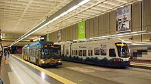 A green bus pulling up to the curb of an underground station, pictured alongside a white train heading in the opposite direction