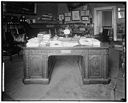 A black and white image of the Resolute desk in a room with dark walls, low bookcases, and memorabilia displayed on the walls. The desk is cluttered with stacks of paper, objects, and a large flower.