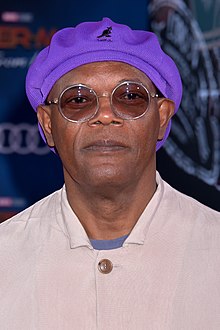 An African-American man is at the centre of the image looking at the camera. He is wearing a purple cap, glasses, and an off-white coat.