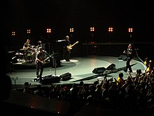 Un escenario colorido y redondo dentro de una arena, con los cuatro miembros de la banda tocando sus instrumentos.